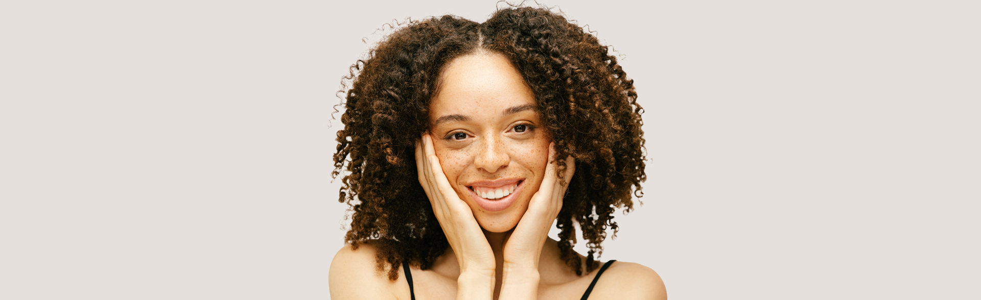 Porträt einer lächelnden Frau mit lockigem Haar und Sommersprossen, die ihr Gesicht mit beiden Händen sanft berührt, vor hellem, neutralem Hintergrund.